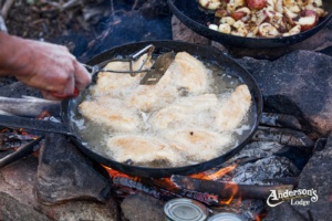 Walleye is cooked in a pan over a fire on the shore of a lake
